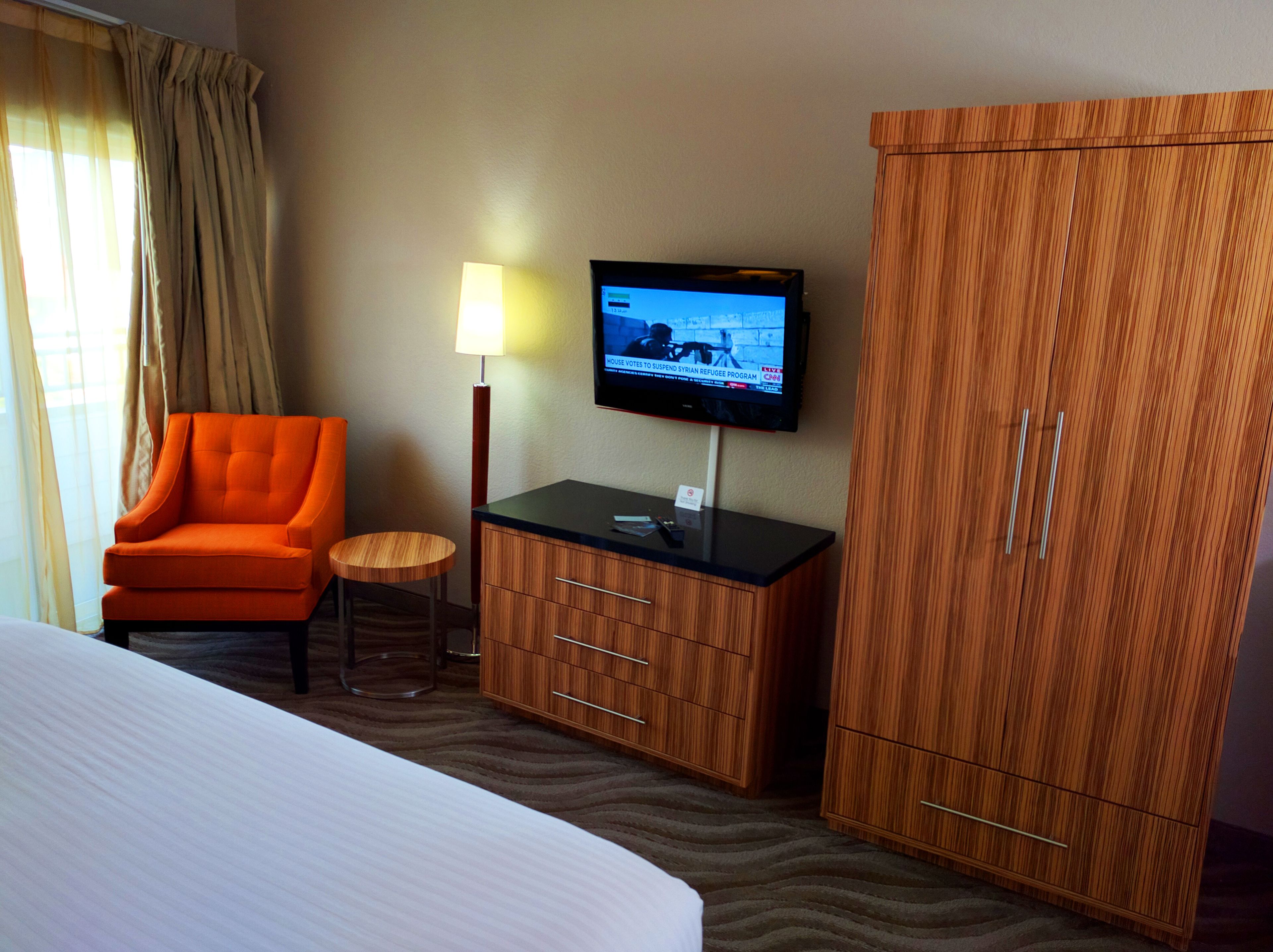 Ein Hotelzimmer mit einem Bett, einem orangefarbenen Sessel, einem kleinen runden Tisch, einem an der Wand befestigten Fernseher und Holzschränken.