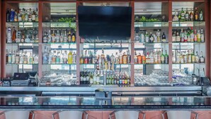 A bar area with shelves of liquor bottles, a TV screen, and a bar counter with glasses and a cash register.