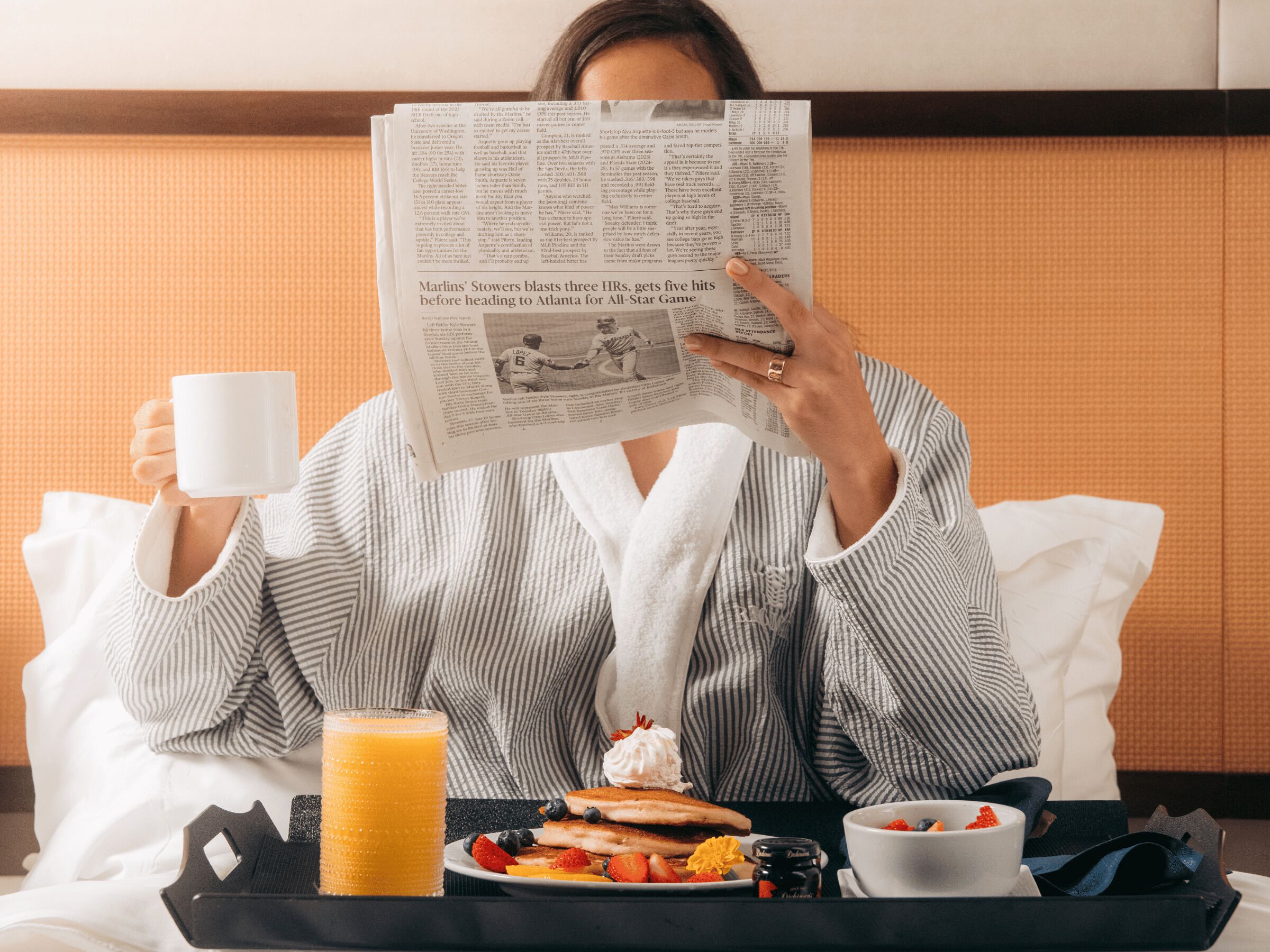 Une personne est assise dans une chambre d’hôtel, en train de lire un journal, avec un plateau de petit-déjeuner devant elle, comprenant des crêpes, des fruits et un verre de jus d’orange.