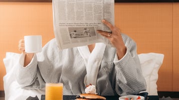 Une personne est assise dans une chambre d’hôtel, en train de lire un journal, avec un plateau de petit-déjeuner devant elle, comprenant des crêpes, des fruits et un verre de jus d’orange.