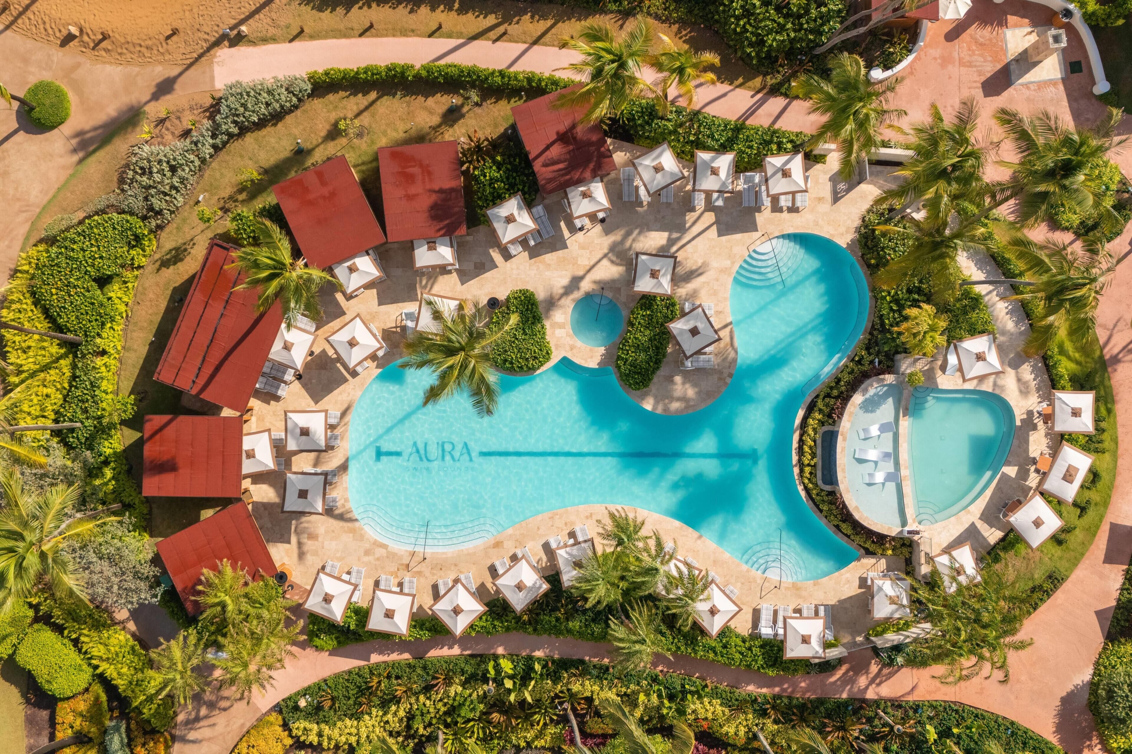 Vista aérea de un complejo turístico con una gran piscina, varias zonas de descanso y palmeras.