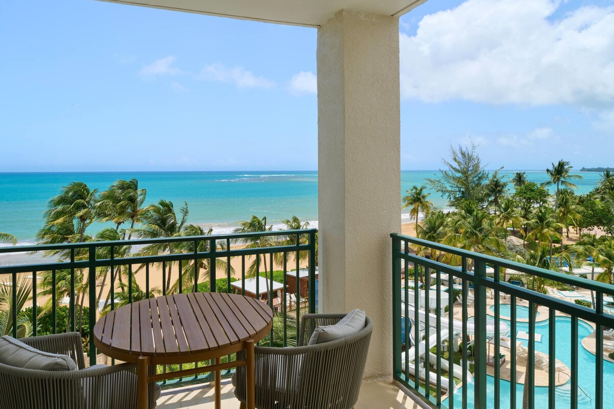 A balcony with a view of the beach and ocean, a pool area, and outdoor seating.