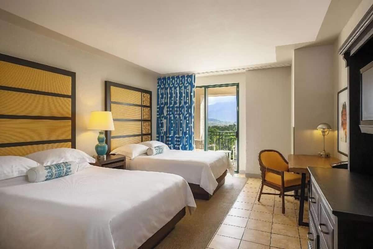 A hotel room with two beds, a desk, a chair, and a balcony with a view.