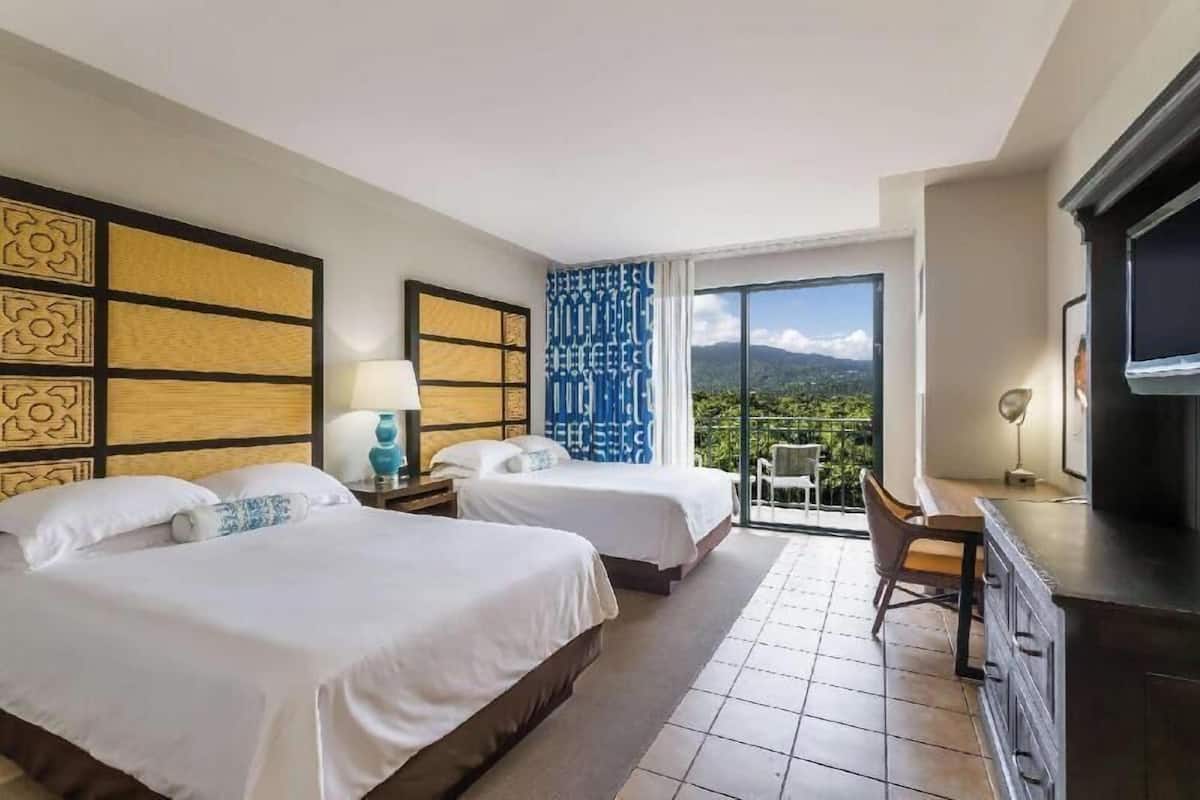 A hotel room with two beds, a desk, a TV, and a balcony with a mountain view.