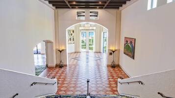 Un escalier mène à un espace vaste doté d’un sol à carreaux, d’un plafond avec des poutres apparentes et d’une vue sur l’extérieur grâce à une grande porte.
