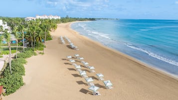 A beach with rows of sun loungers and umbrellas, a clear blue ocean, and a resort building in the background.