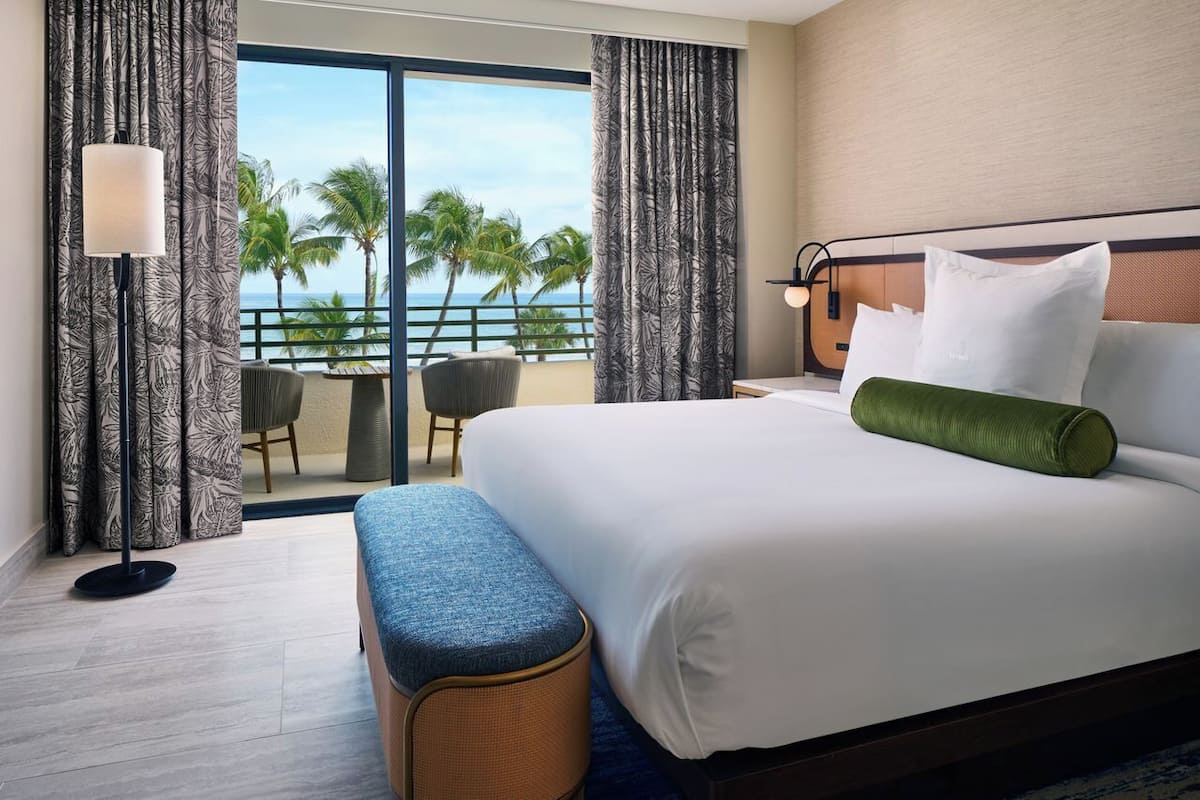 A hotel room with a bed, a bench, a nightstand, a lamp, a view of palm trees and the ocean.