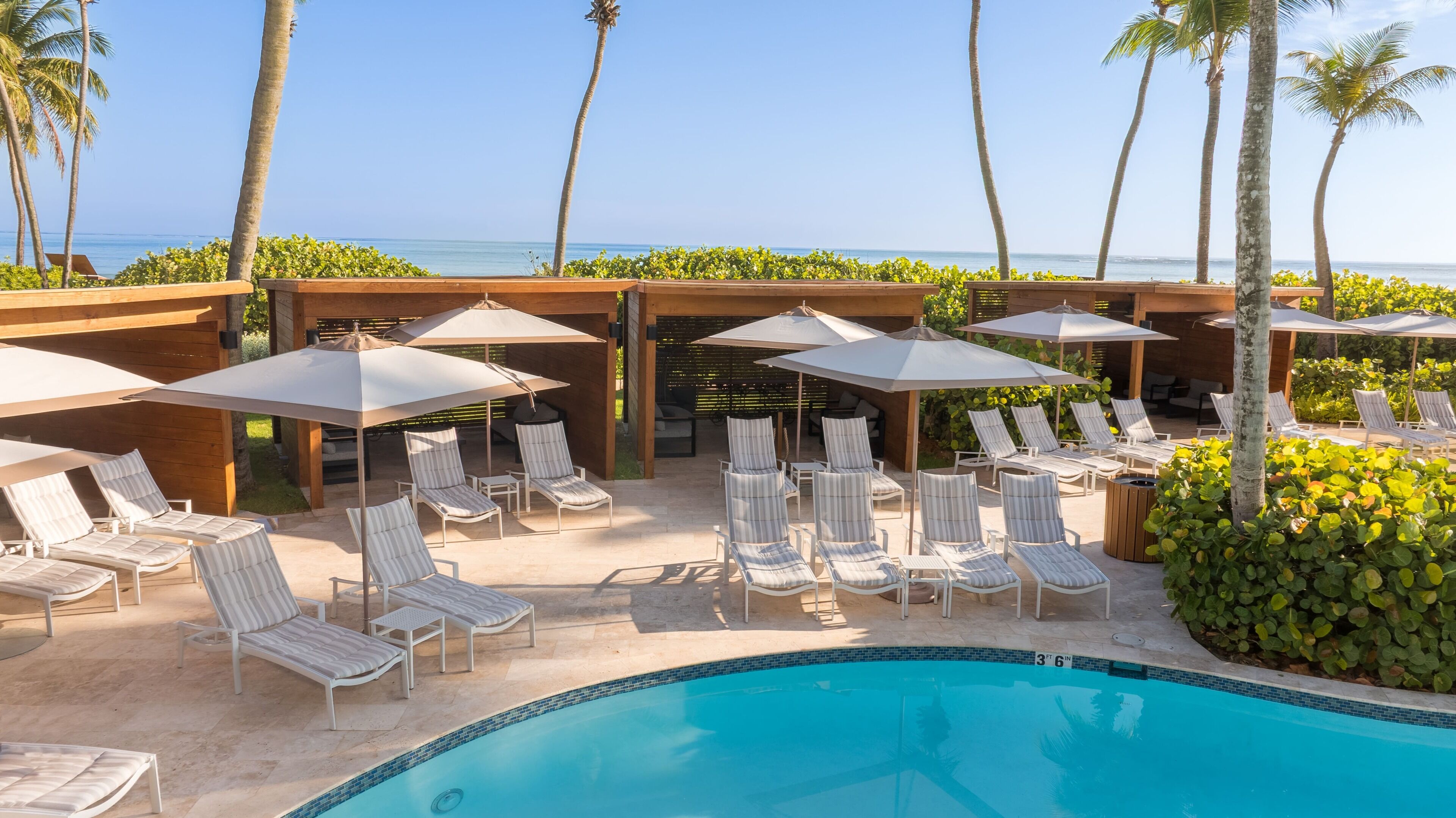 Un espace aménagé au bord de la piscine, avec des chaises longues et des parasols, offrant une vue dégagée sur l’océan, et agrémenté d’une structure en bois formant une pergola.
