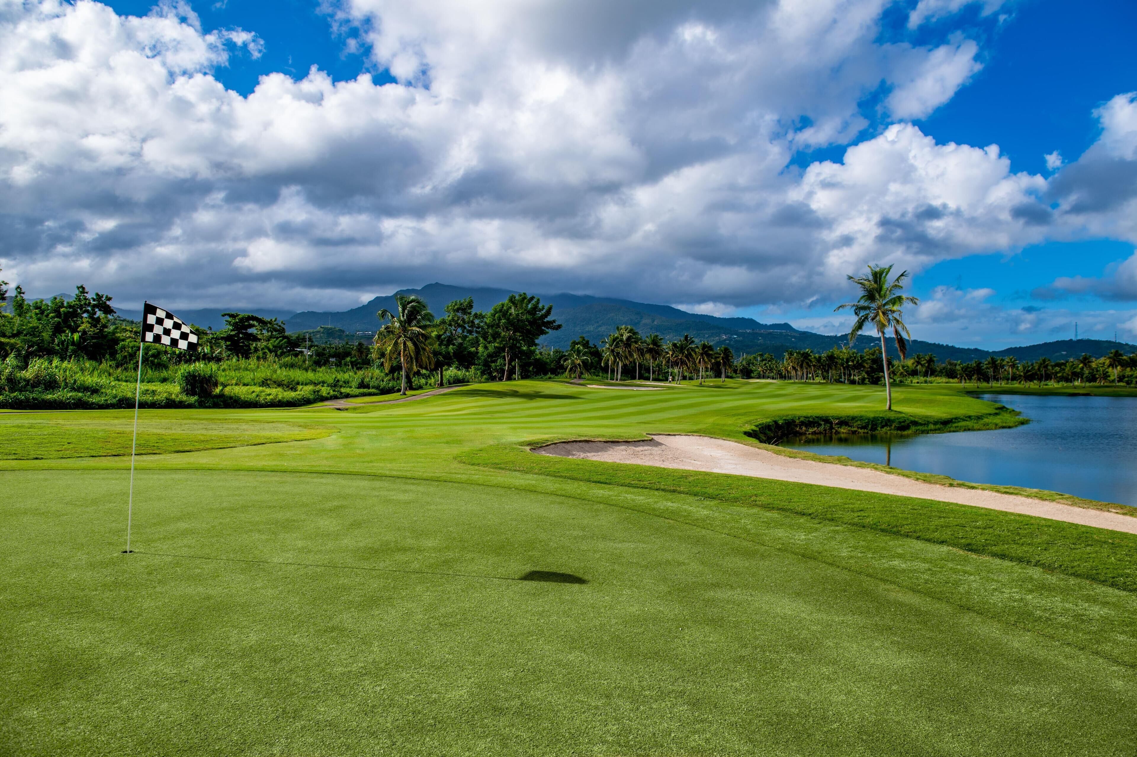 Un parcours de golf avec un mât de drapeau, un plan d’eau, des palmiers et des montagnes en arrière-plan.