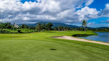 Un parcours de golf avec un mât de drapeau, un plan d’eau, des palmiers et des montagnes en arrière-plan.