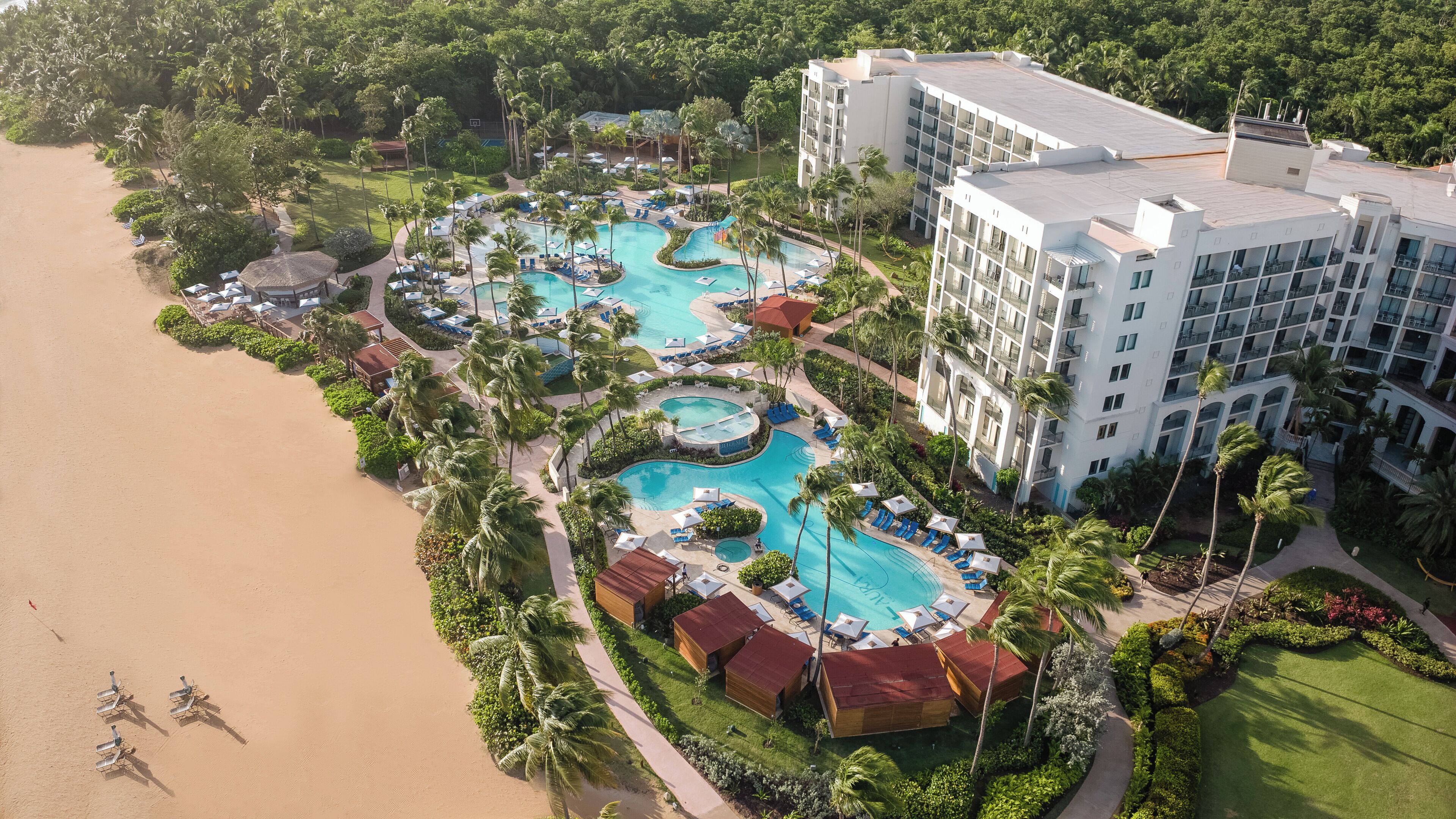 An aerial view of a resort with multiple swimming pools, beachfront, and a large hotel building.