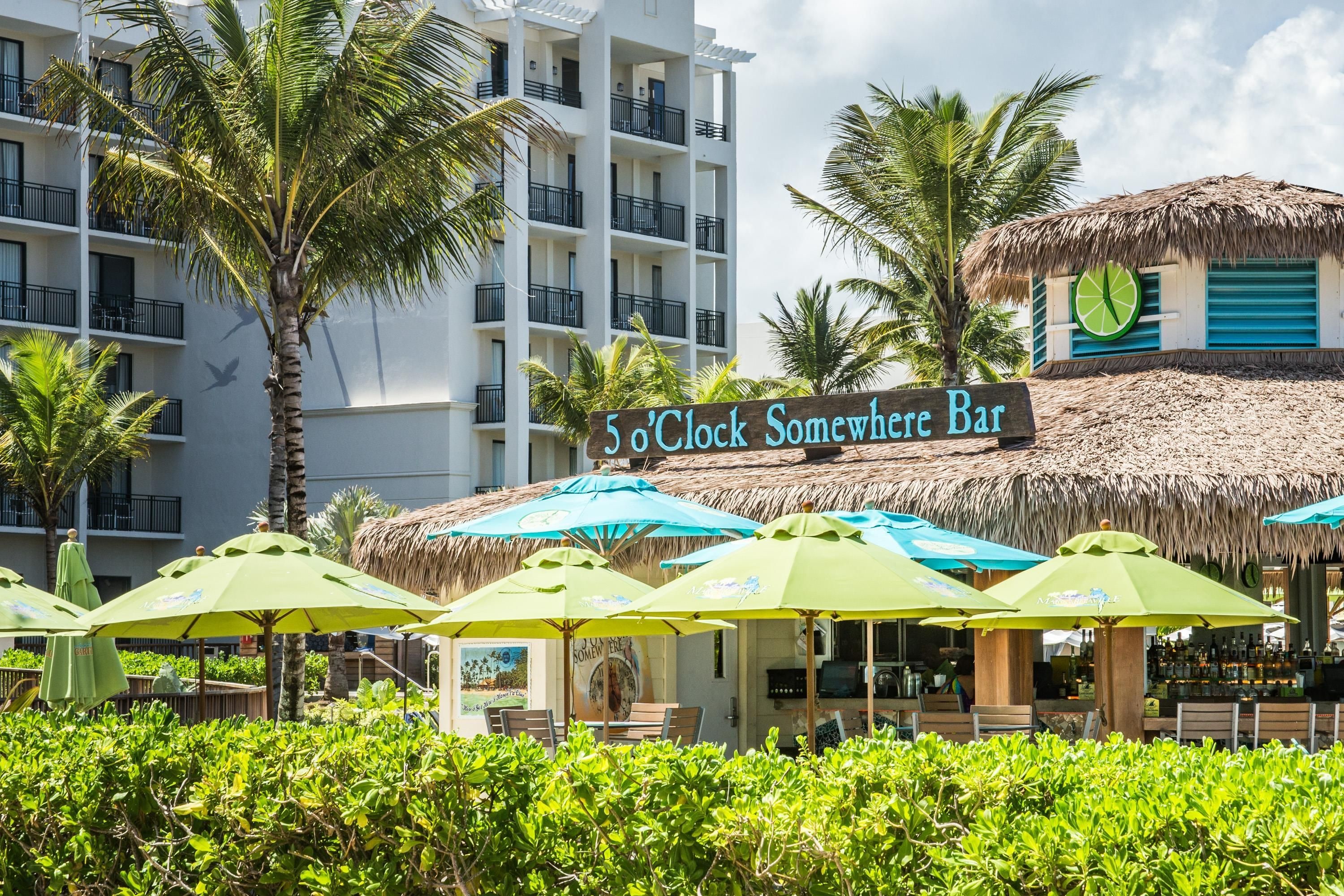 Un bar d’hôtel avec une enseigne indiquant « 5’0 Clock Somewhere Bar » et des parasols en chaume.