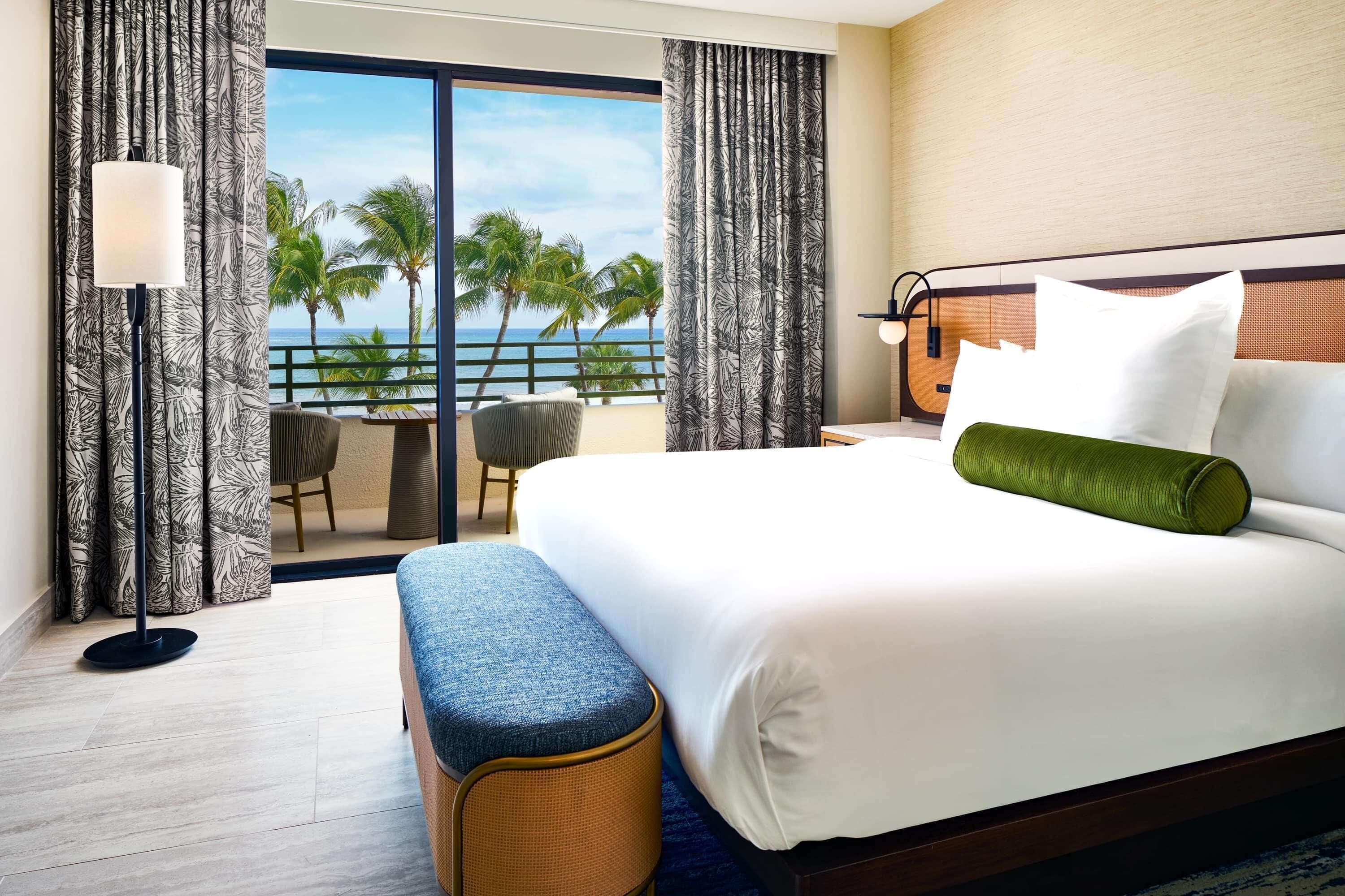 Habitación de hotel con una cama grande, un balcón con vistas a palmeras y al mar, una silla y una lámpara.