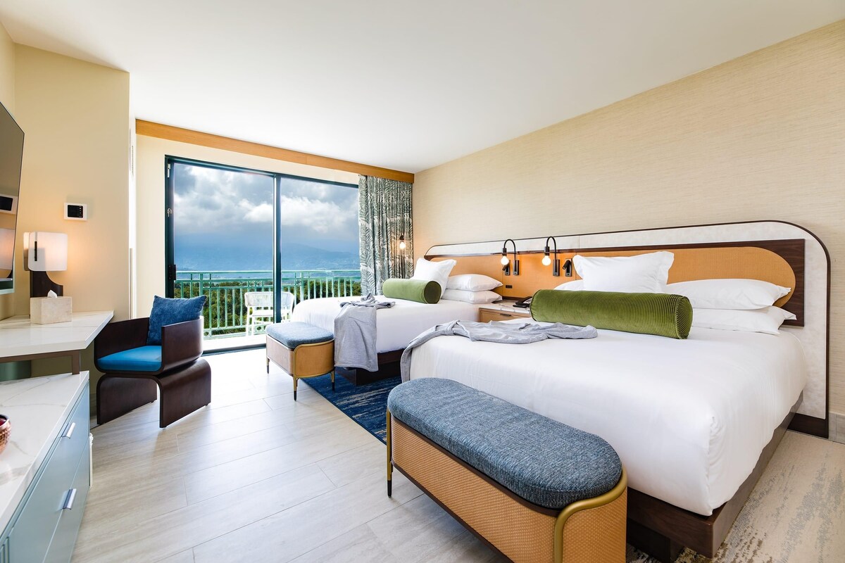A hotel room with a large bed, a desk, a chair, a bench, and a balcony with a view.