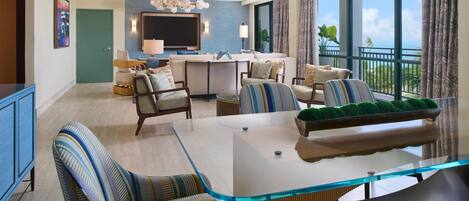 A modern living room with a glass-top dining table, striped upholstered chairs, a ceiling fan, and a view of the ocean through large windows.