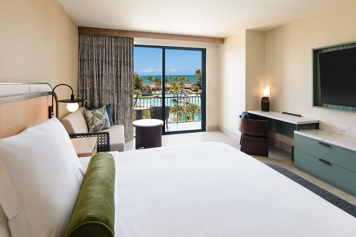 A hotel room with a bed, a desk, a chair, a small table, a TV, and a balcony with a pool view.