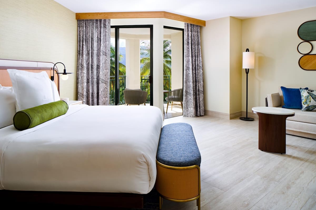 A hotel room with a large bed, a bench, a small table, a sofa, and a view of the outdoors.