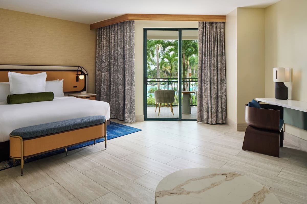 A hotel room with a bed, a desk, a chair, and a view of a pool and palm trees.