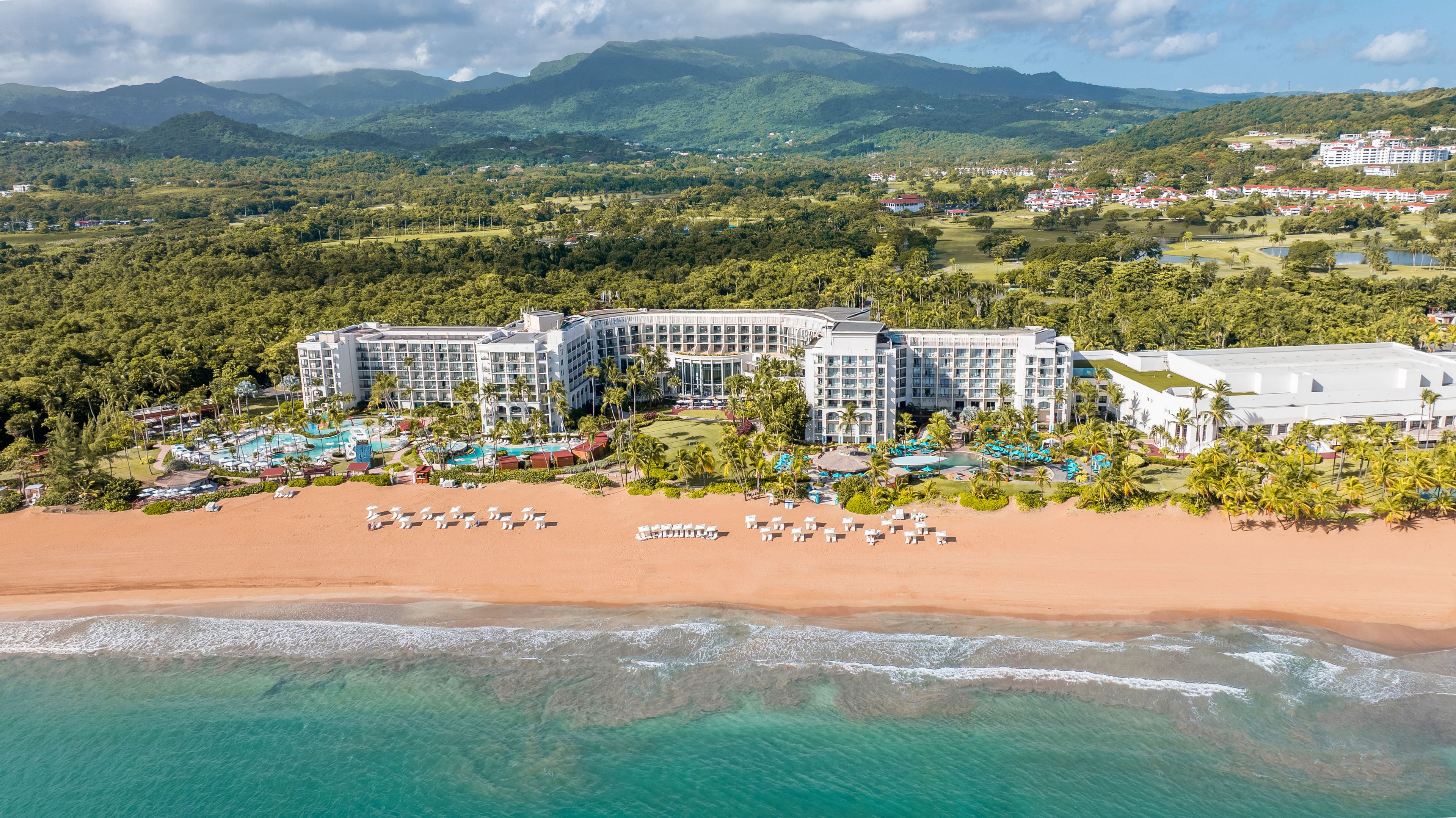 A resort with multiple buildings, a beachfront, and a mountainous backdrop.