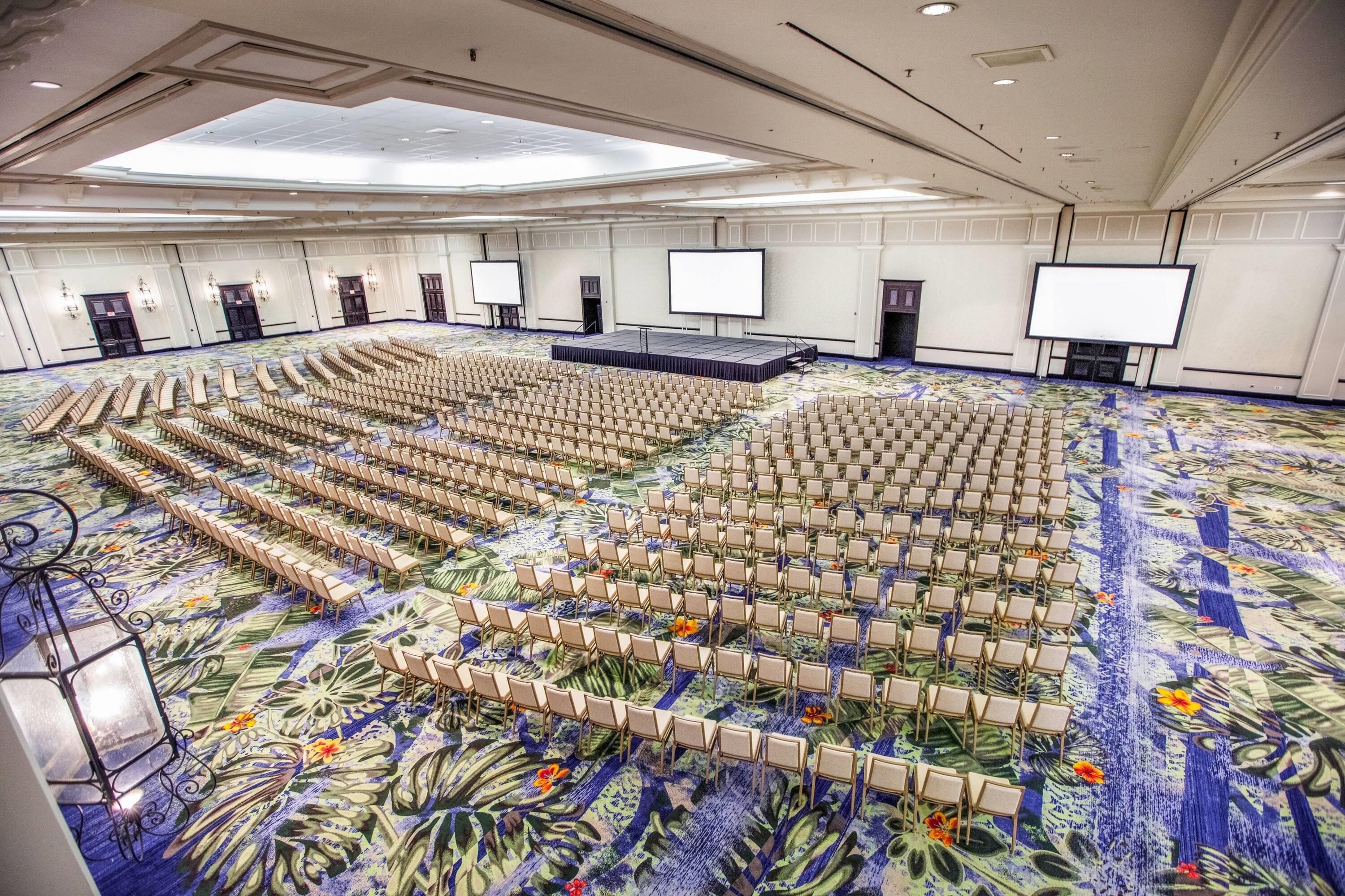 Un salón de conferencias amplio con filas de sillas, un escenario y una pantalla grande.