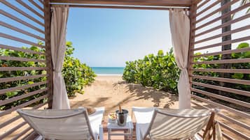 A beach view from a covered seating area with two chairs and a table, a bottle of champagne, and a bowl of fruit.