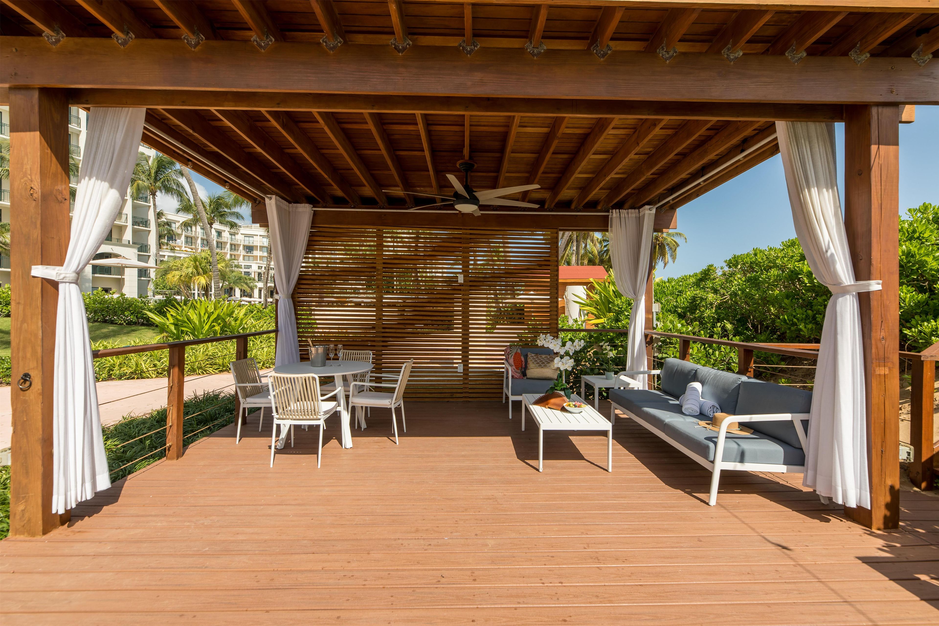 Un espace extérieur abrité, aménagé avec un plancher en bois, du mobilier blanc et offrant une vue sur un bâtiment d’hôtel en arrière-plan.