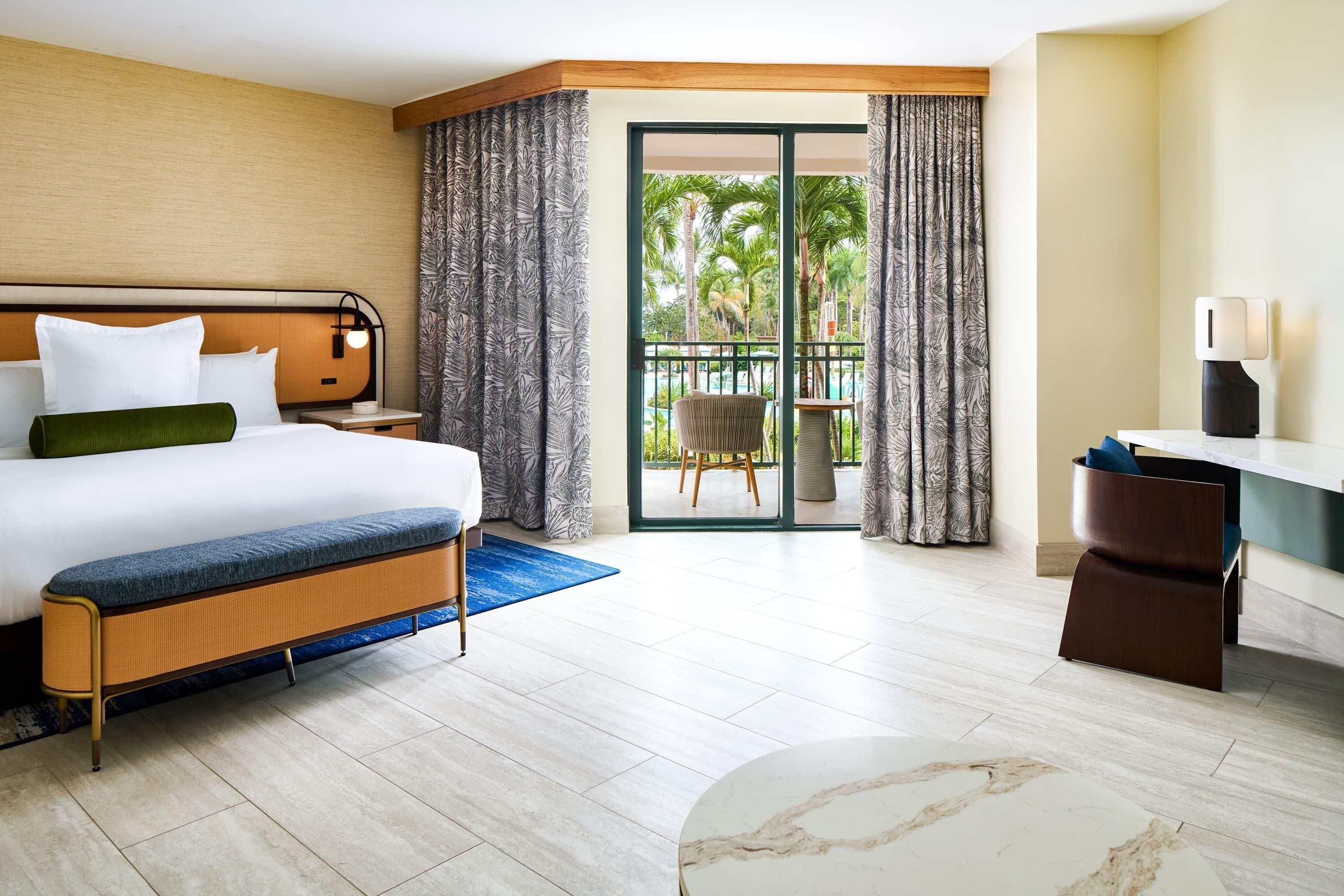 Habitación de hotel con cama, escritorio, silla, balcón y vistas a palmeras, además de una ventana con cortinas.