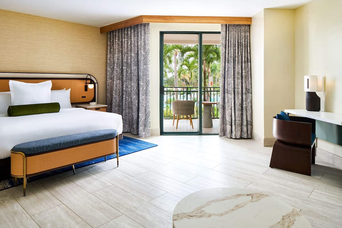 Habitación de hotel con cama, escritorio, silla, balcón y vistas a palmeras, además de una ventana con cortinas.