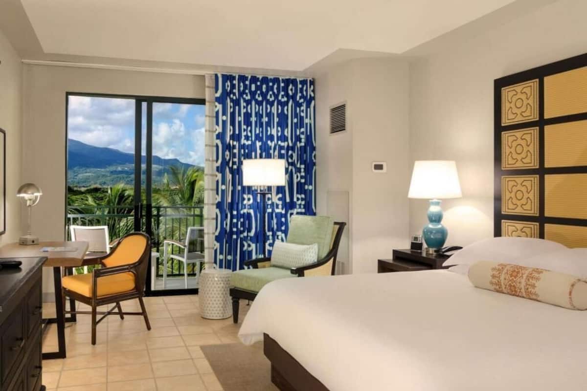 A hotel room with a bed, two chairs, a desk with a computer, a lamp, and a view of the outdoors.