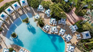 An aerial view of a resort with a large pool, surrounded by lounge areas with umbrellas and chairs, and palm trees.