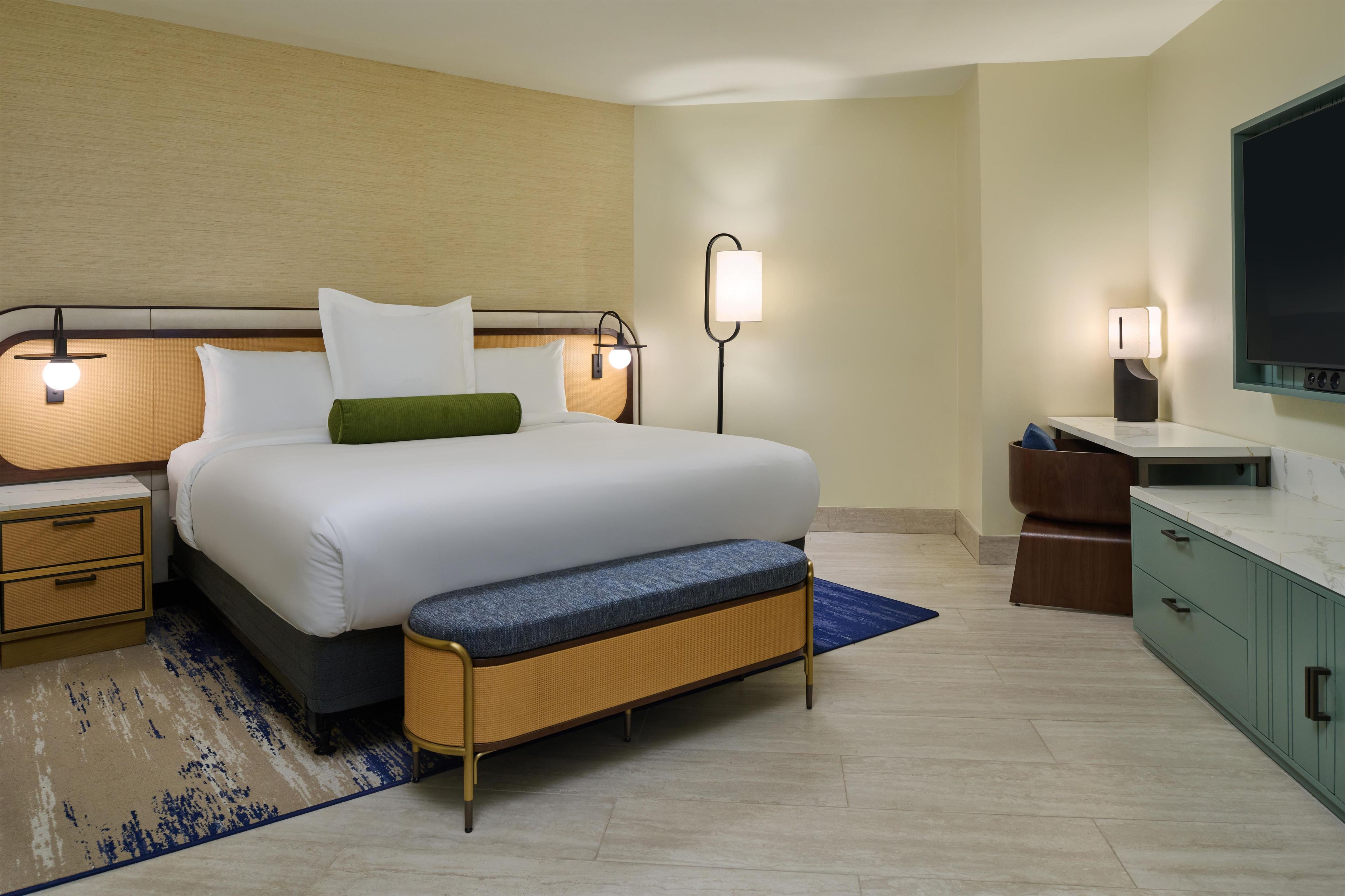 Une chambre d’hôtel comprenant un lit, des tables de chevet, un banc, une télévision et un bureau avec une chaise.