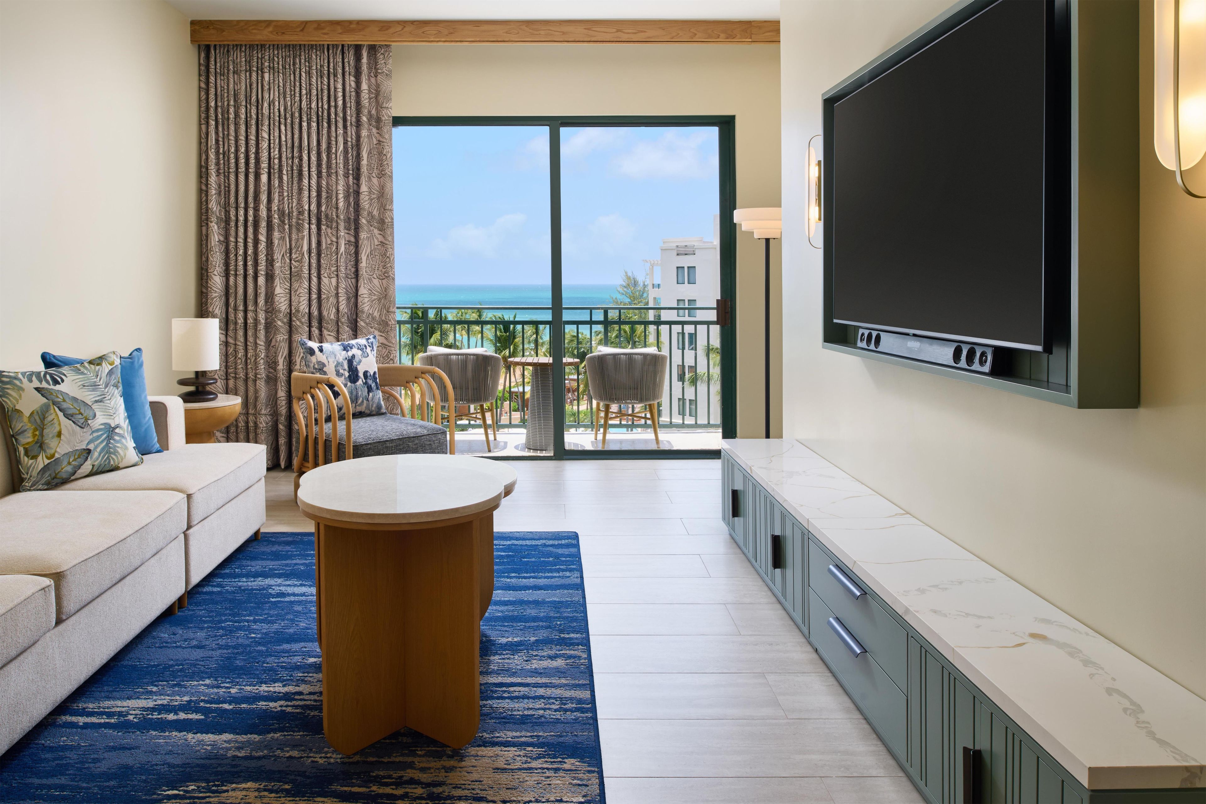 Una sala de estar con sofá, una mesa de centro redonda, un televisor montado en la pared y un balcón con vista al mar.