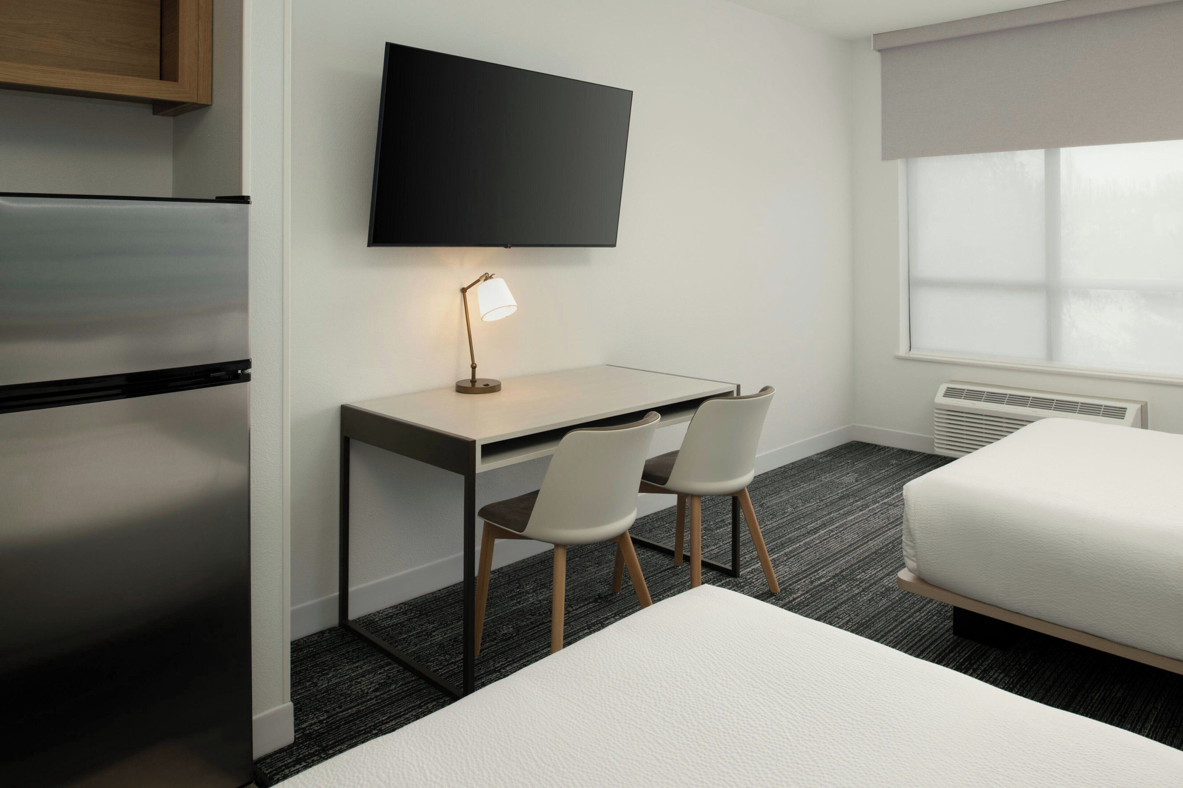Habitación de hotel con cama, escritorio con dos sillas, televisor de pantalla plana, refrigerador y ventana con persianas.