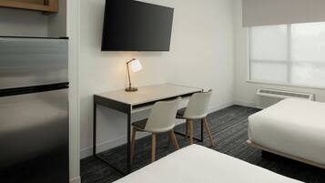 Habitación de hotel con cama, escritorio con dos sillas, televisor de pantalla plana, refrigerador y ventana con persianas.