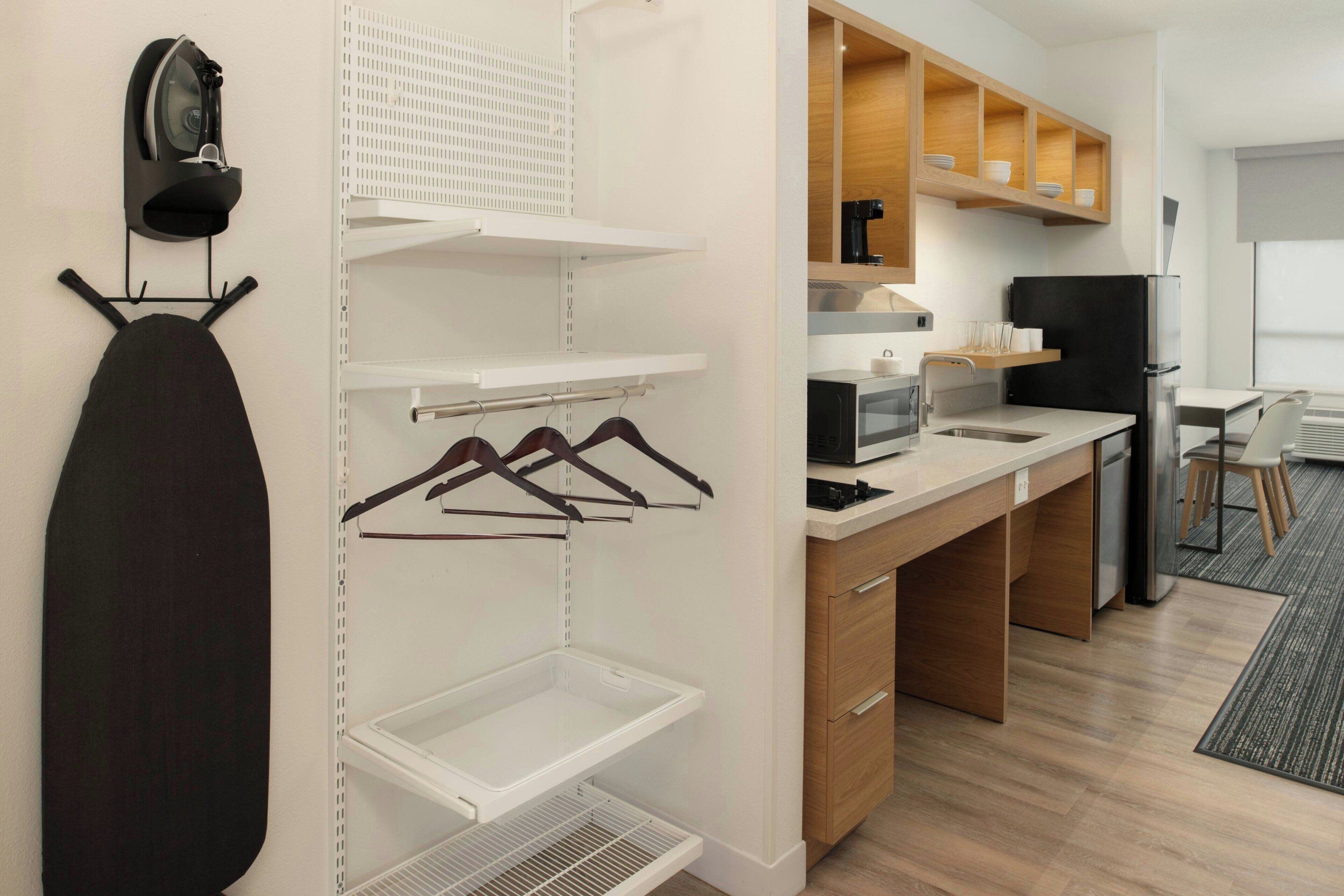 A modern kitchen with wooden cabinets, a black refrigerator, and a microwave. There is a hanging iron and a drying rack on the wall.
