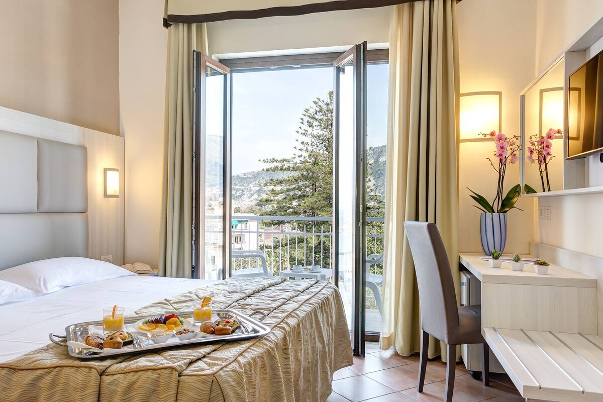 A hotel room with a bed, breakfast tray, desk, chair, and a balcony with a view of the city.