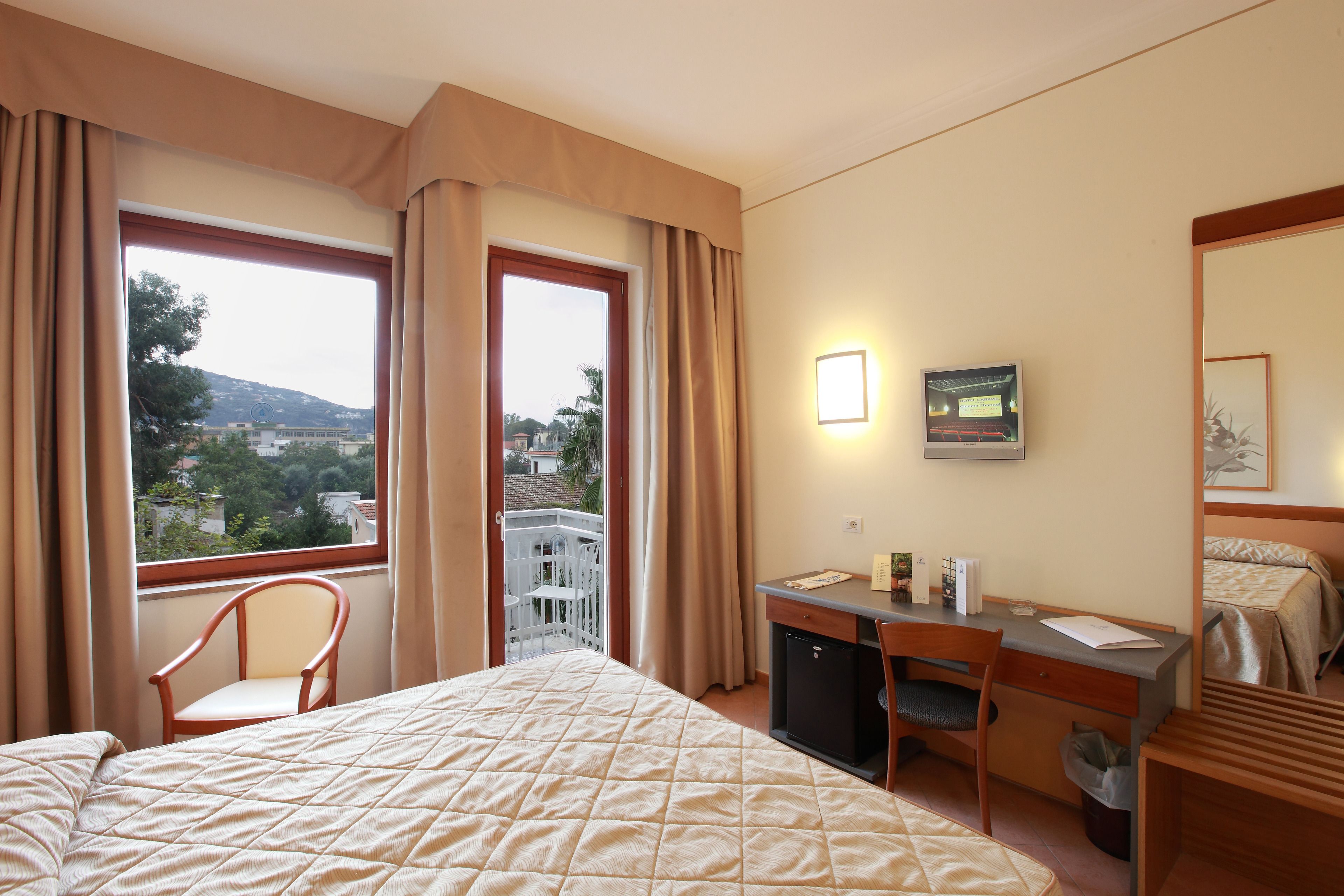 Quarto de hotel com cama, escrivaninha, cadeira e vista da cidade através de amplas janelas.