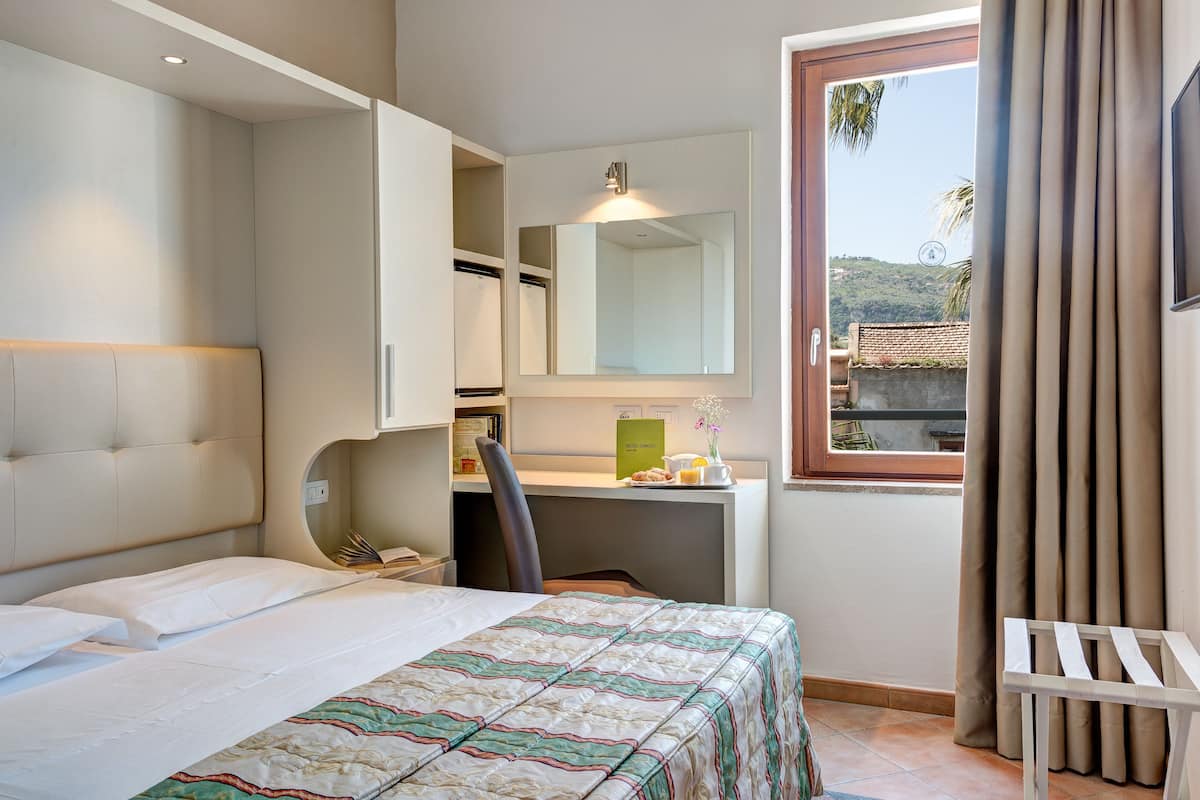 A hotel room with a bed, a desk with a chair, a mirror, and a window with curtains.