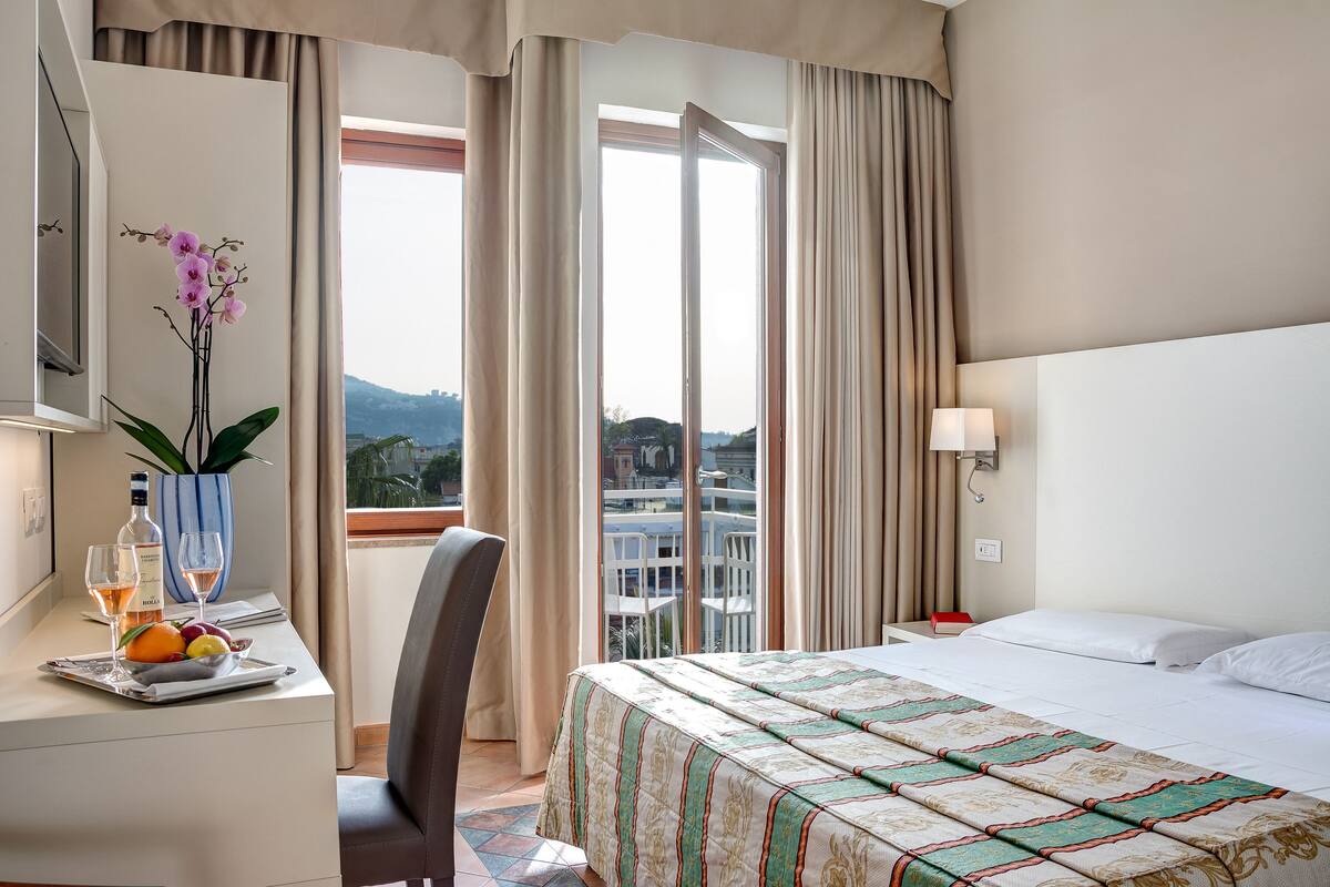A hotel room with a bed, a desk with a vase of flowers, a fruit tray, and a view of the city through the window.