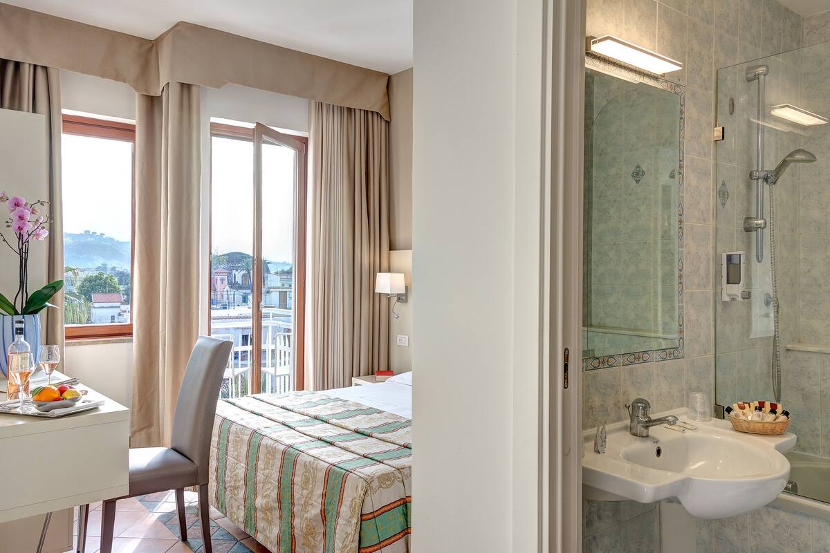 A hotel room with a bed, a desk with a vase and fruit, a view of the city, and a bathroom with a sink and shower.