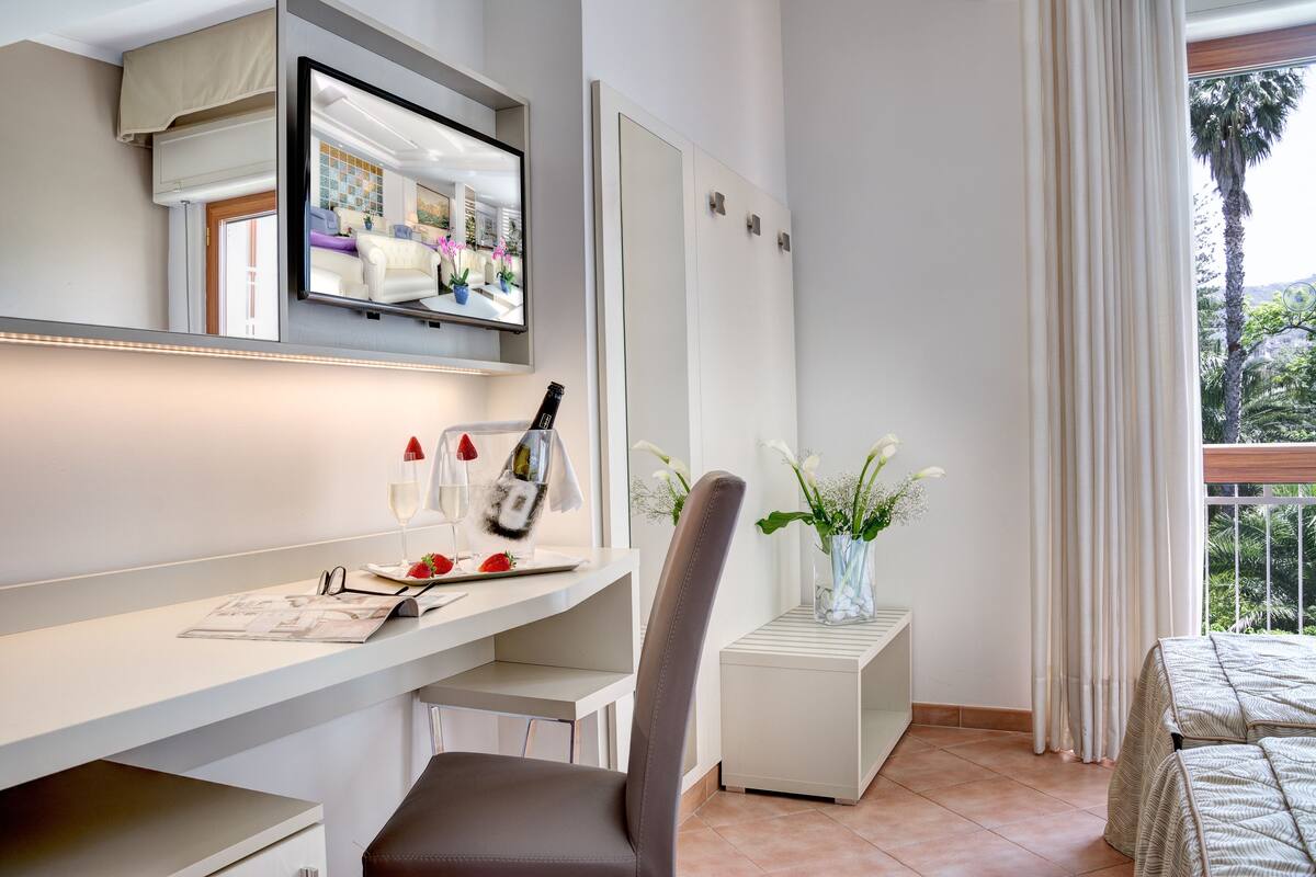 A modern hotel room with a large TV on the wall, a desk with a laptop, a chair, a small side table with a vase of flowers, and a bed with a view of the outdoors.