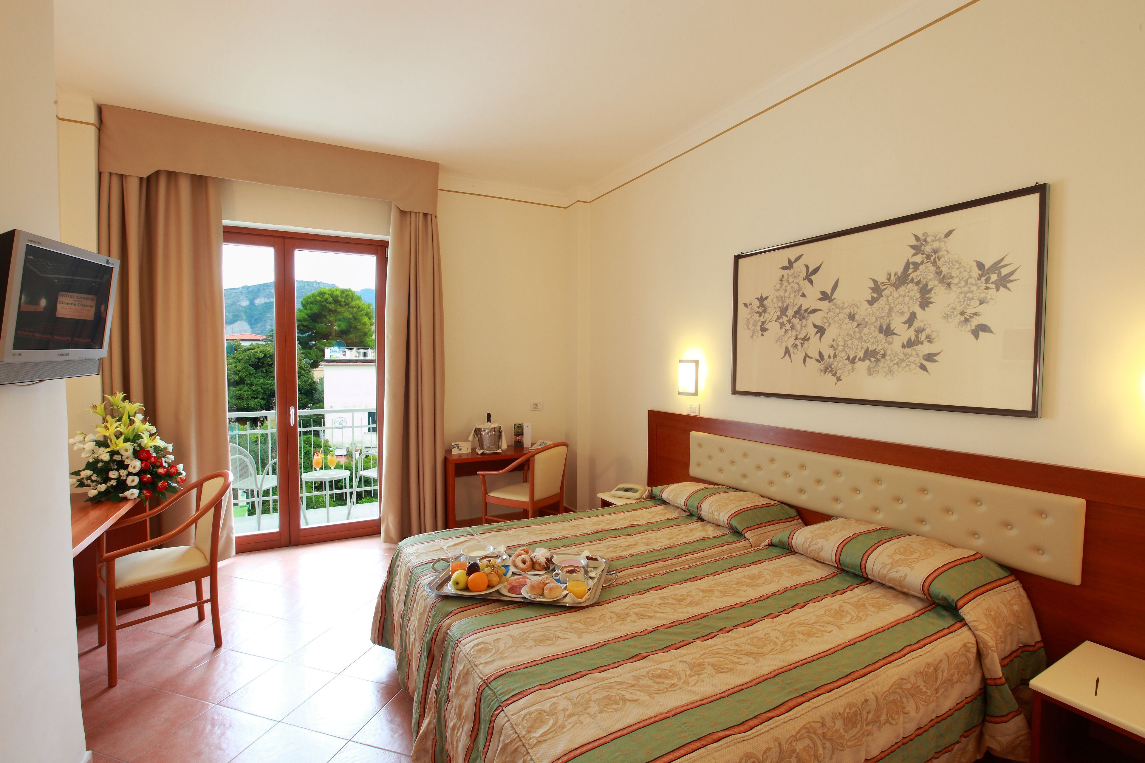 A hotel room with a bed, a desk with a chair, a small table with a lamp, and a balcony with curtains.
