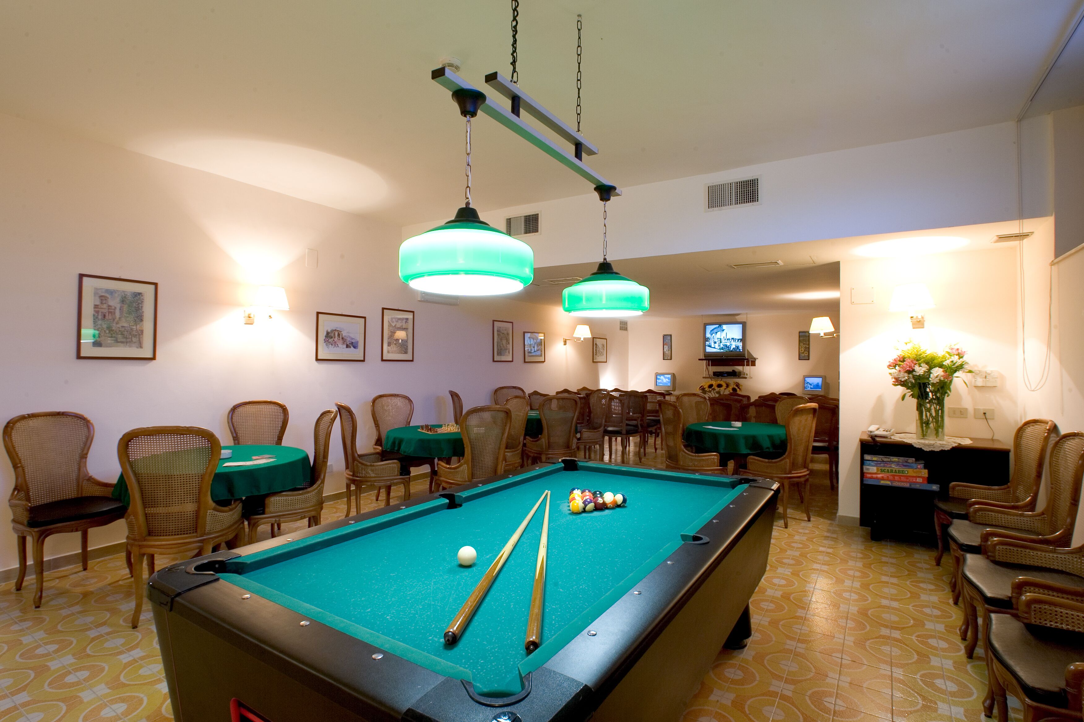 A pool table with billiard balls, surrounded by wicker chairs and tables, with overhead lighting and framed pictures on the walls.