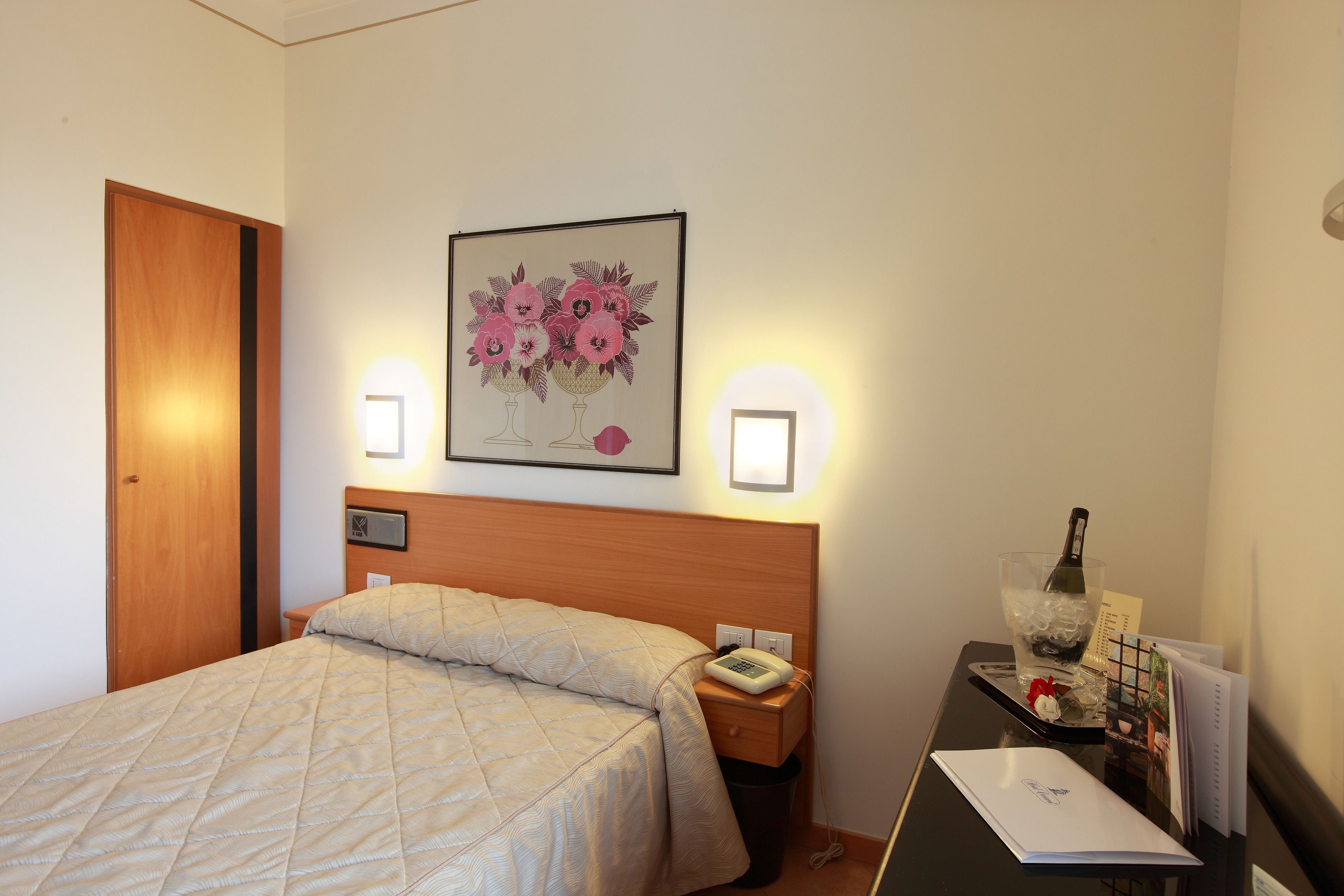 Ein Hotelzimmer mit einem Bett, einem Nachttisch mit Telefon, einem gerahmten Bild an der Wand und einem kleinen Tisch mit Flasche und Gläsern.