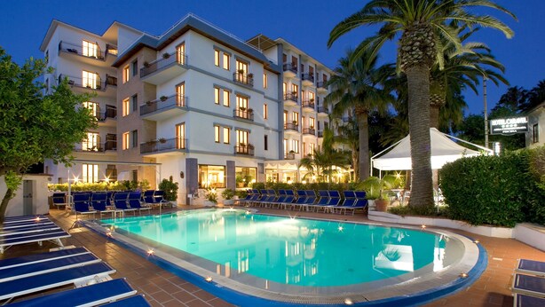 A hotel with a swimming pool, lounge chairs, and palm trees. - Hotel Caravel (Sant'Agnello)