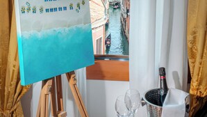 Une vue par la fenêtre sur un canal, avec des bâtiments, des rideaux, un chevalet sur lequel est posée une toile, et un plateau avec des verres et une bouteille.