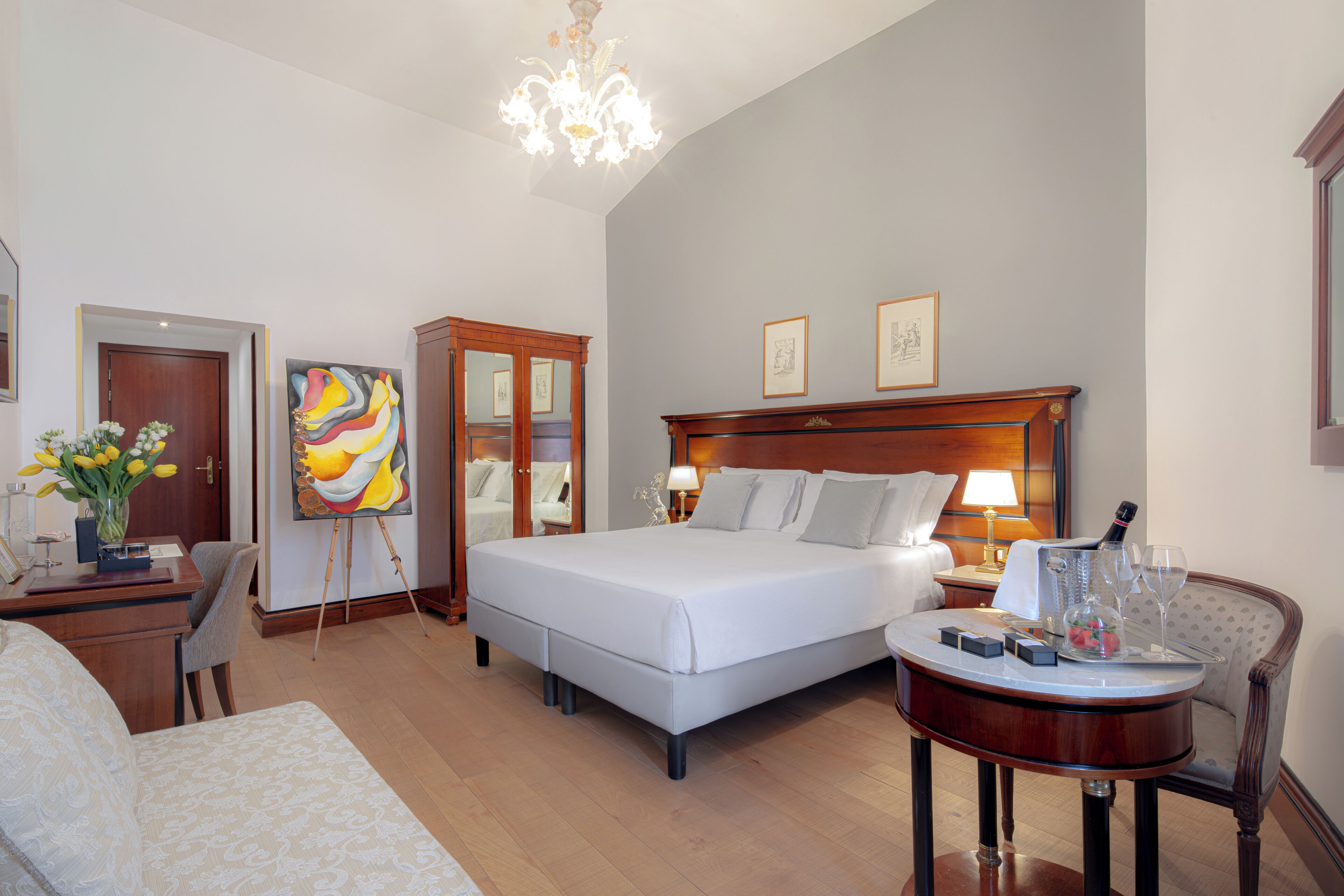 A hotel room with a large bed, a desk with a chair, a round table with a bottle and glasses, and artwork on the walls.