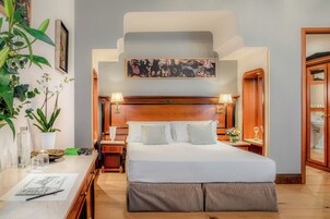 Quarto de hotel com uma cama grande, cabeceira de madeira, duas mesas de cabeceira com luminárias, um criado-mudo e um vaso de planta, além de uma obra de arte emoldurada acima da cama.