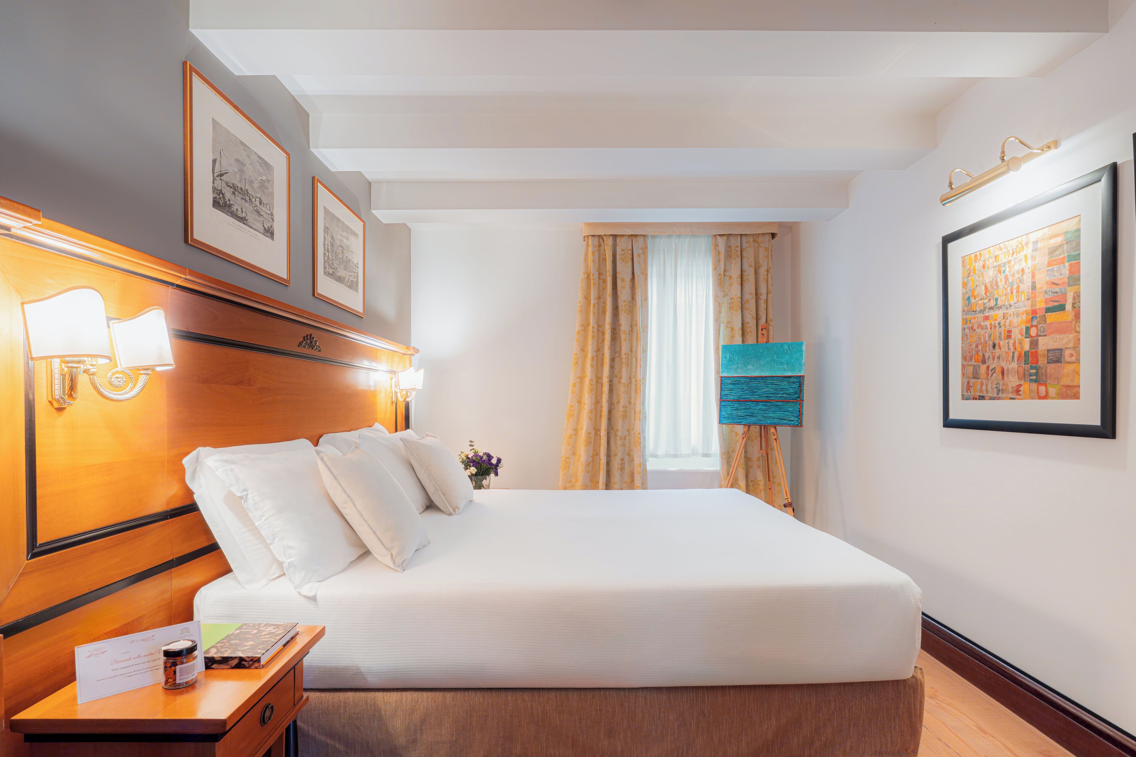 A hotel room with a neatly made bed, a nightstand with a lamp, framed pictures on the wall, and a window with curtains.