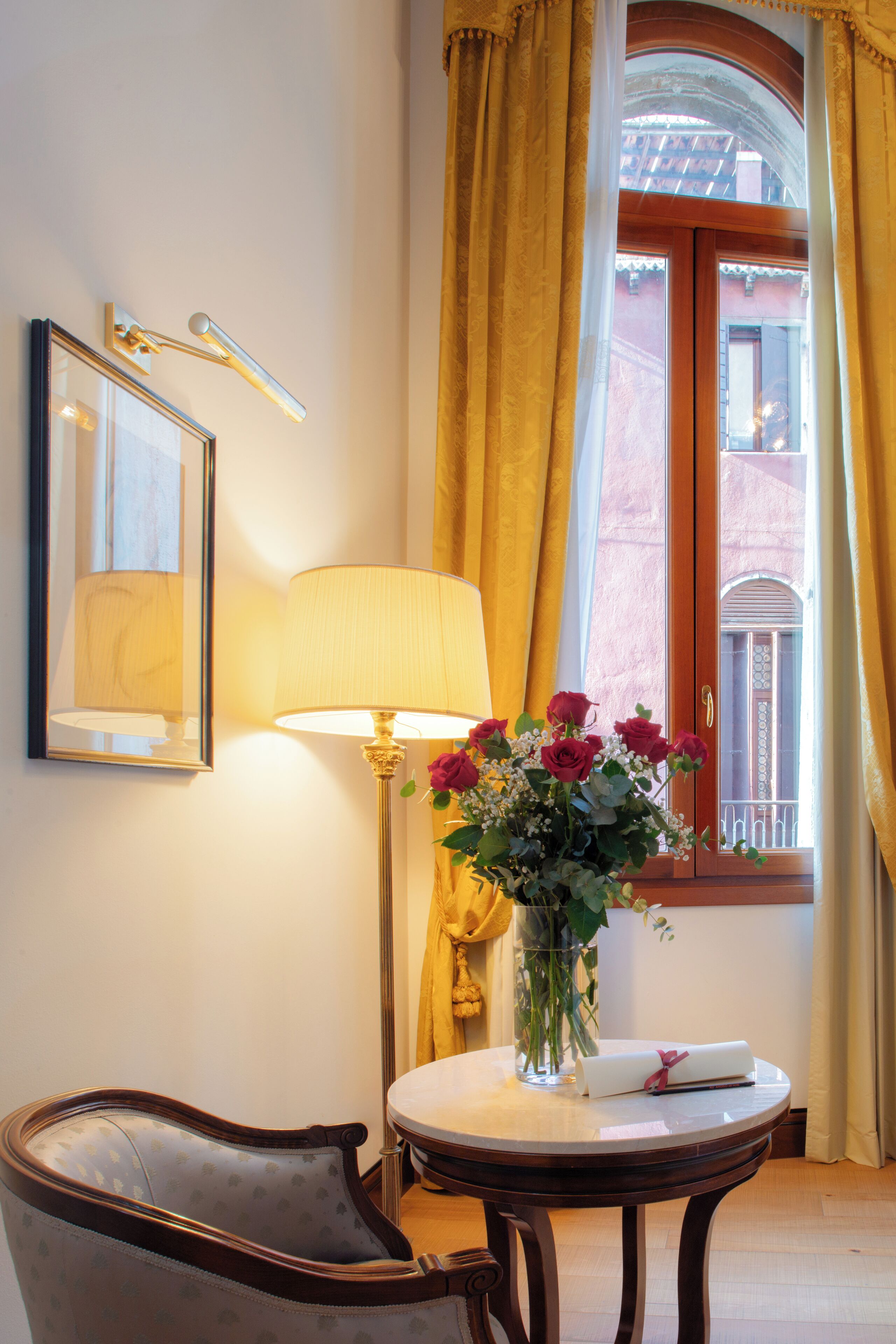 A room with a round table, a chair, a lamp, a vase with flowers, a framed picture, and a window with curtains.